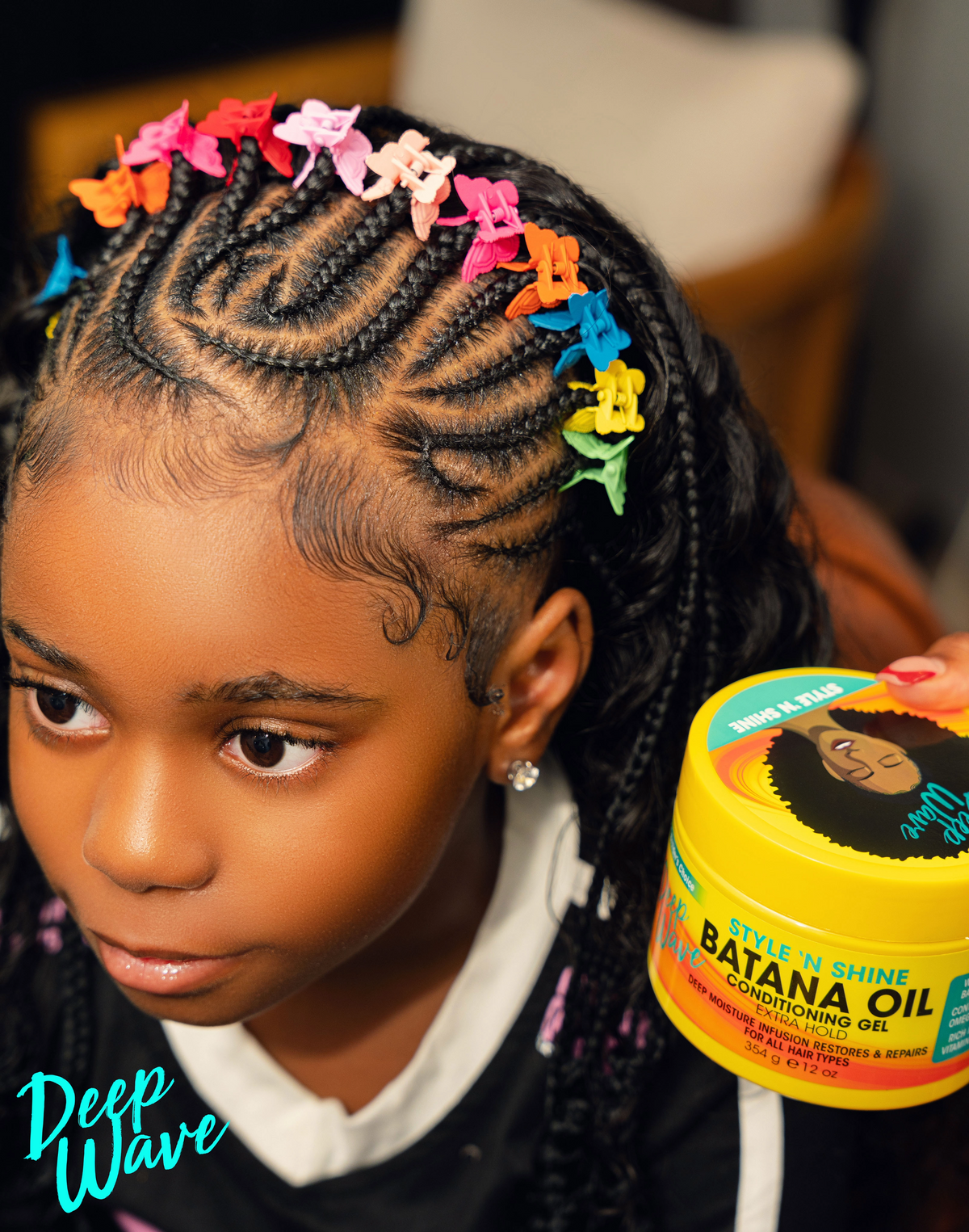A close-up lifestyle shot of a young girl with braided hair decorated with colorful butterfly clips, holding a yellow tub of Deep Wave Batana Oil Conditioning Gel.