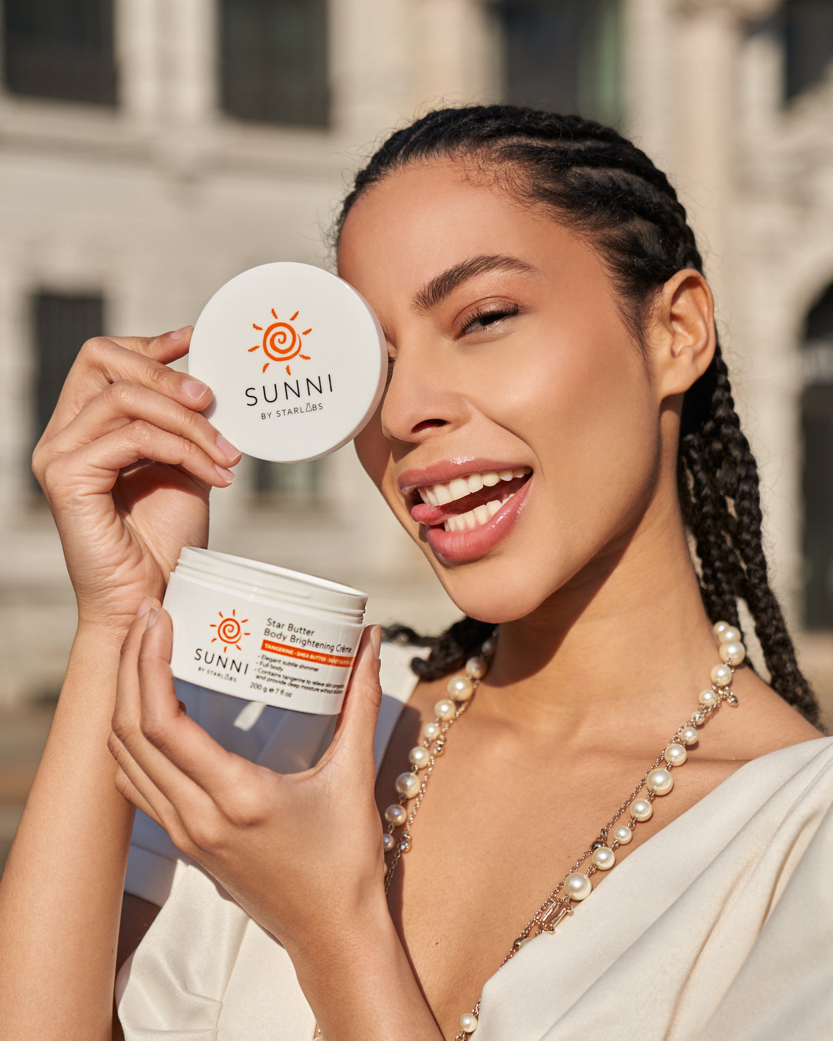 Woman holding a white skincare jar and lid labeled SUNNI, smiling outdoors with braided hair visible