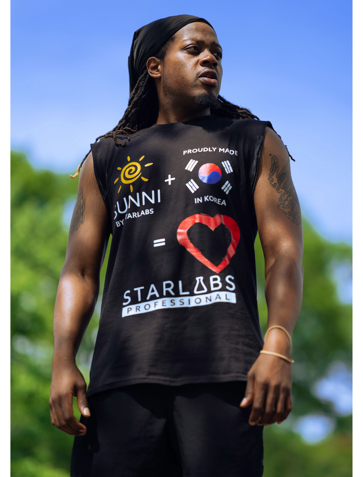A man wearing a black sleeveless muscle tank top with a colorful graphic print featuring the text 'SUNNI x STARLABS PROFESSIONAL' and a red heart design, photographed outdoors against a blurred blue sky and green foliage background.