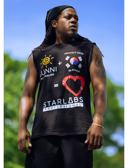 A man wearing a black sleeveless muscle tank top with a colorful graphic print featuring the text 'SUNNI x STARLABS PROFESSIONAL' and a red heart design, photographed outdoors against a blurred blue sky and green foliage background.