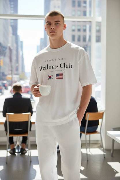 Man in a white t-shirt with Starlabs Wellness Club text holding a mug in a modern office setting