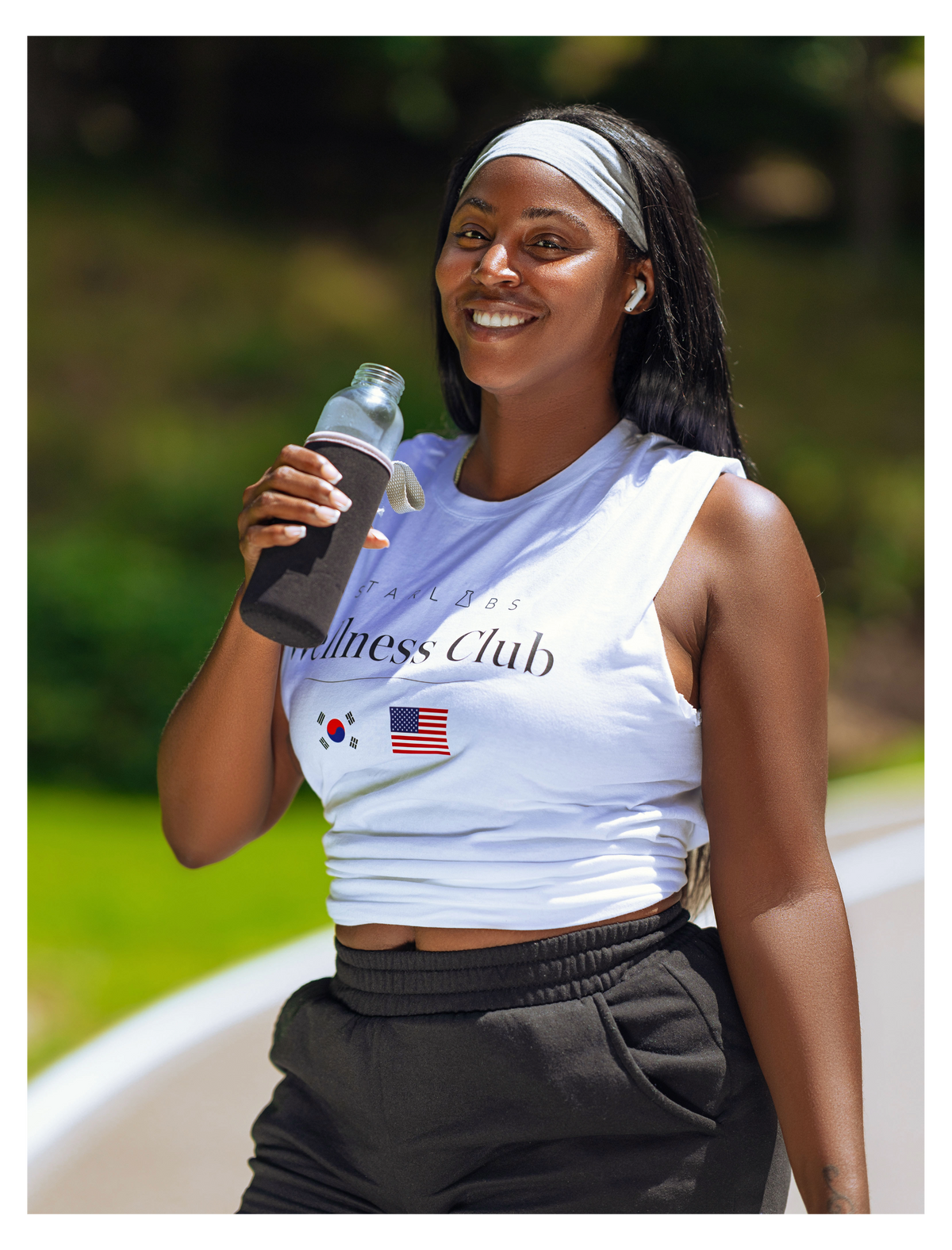 A woman outdoors wearing a white sleeveless athletic tank top with 'Wellness Club' text and small South Korean and American flag graphics, paired with dark sweatpants and holding a water bottle.