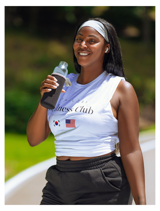 A woman outdoors wearing a white sleeveless athletic tank top with 'Wellness Club' text and small South Korean and American flag graphics, paired with dark sweatpants and holding a water bottle.