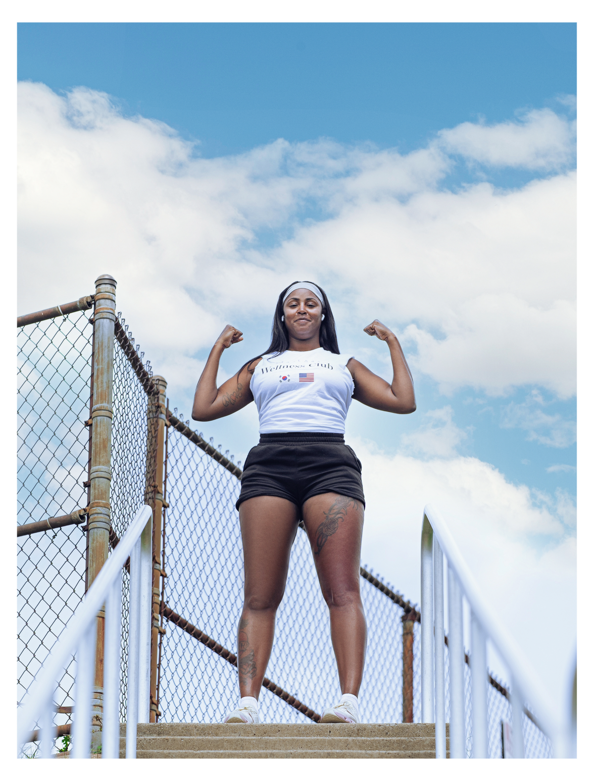 A woman stands on outdoor concrete steps under a blue sky with clouds, flexing her biceps. She is wearing a white athletic 'Star Labs Wellness Club' with a small American flag and Korean flag graphic and the text paired with black athletic shorts.