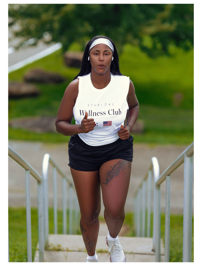 A woman running up outdoor steps wearing a white tank top with 'Wellness Club' printed on the front, black athletic shorts, and white sneakers.