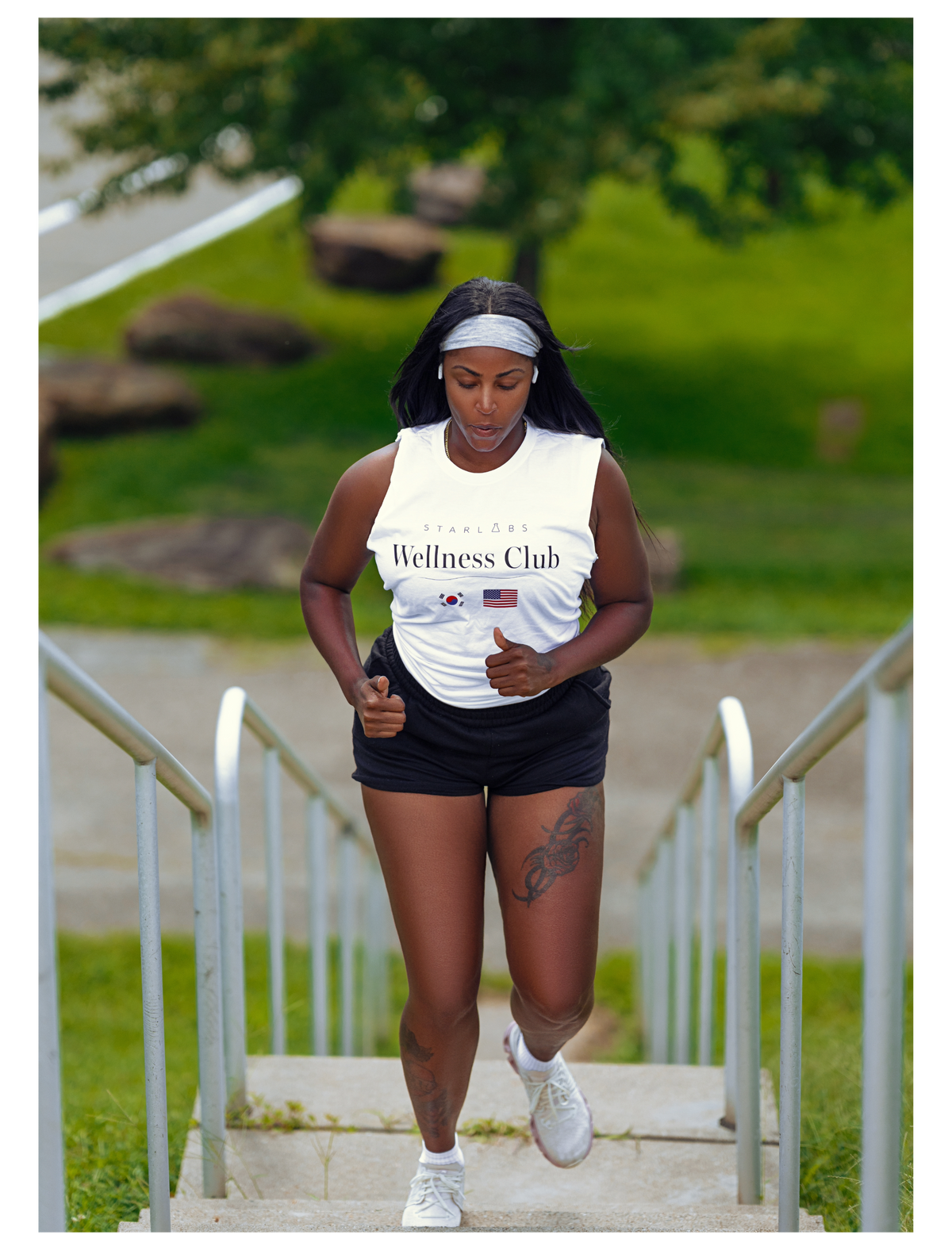 A woman running up outdoor concrete stairs wearing a white tank top with 'Wellness Club' printed on the front, black athletic shorts, and white sneakers.