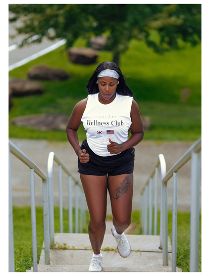 A woman running up outdoor concrete stairs wearing a white tank top with 'Wellness Club' printed on the front, black athletic shorts, and white sneakers.